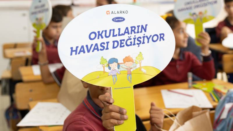 Sınıfta sıralarında oturan öğrenciler, “Okulların Havası Değişiyor” yazılı yuvarlak kartonları tutuyor