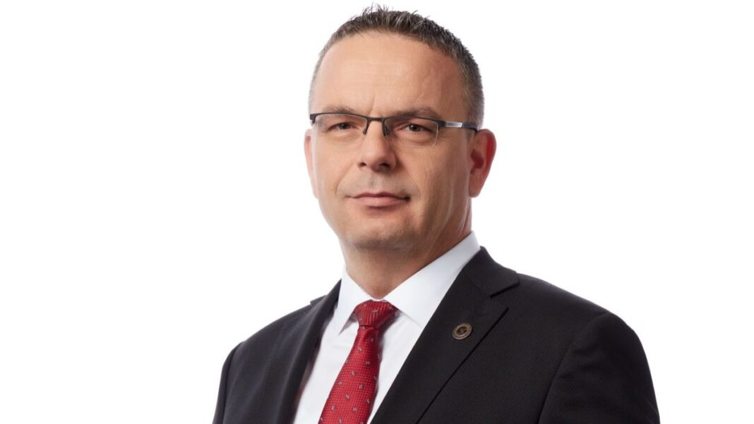Koyu takım elbise ve kırmızı kravatlı, gözlüklü Sezgin Yılmaz’ın beyaz fon önünde portresi