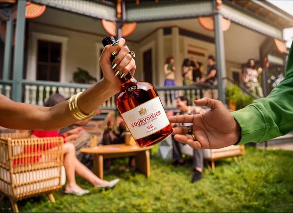 Bahçede düzenlenen bir ev partisinde bir elden diğerine uzatılan Courvoisier şişesi; arka planda verandada sohbet eden konuklar