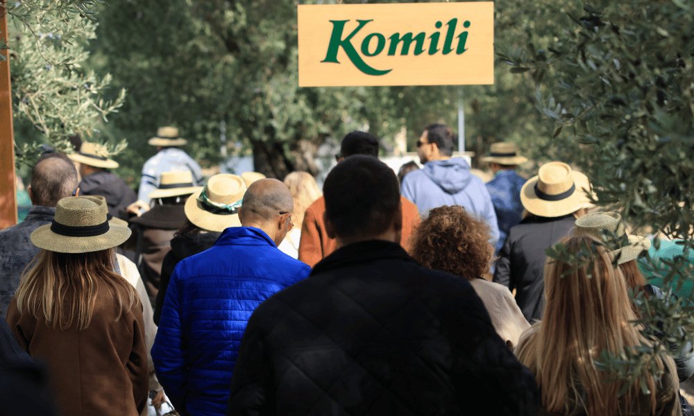 Ayvalık’taki zeytin bahçesinde Komili tabelasının önünde toplanmış, şapkalı misafirlerden oluşan kalabalık