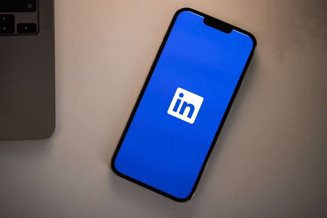 Masa üzerinde, dizüstü bilgisayarın yanında ekranında mavi zemin üzerinde LinkedIn logosu görünen akıllı telefon