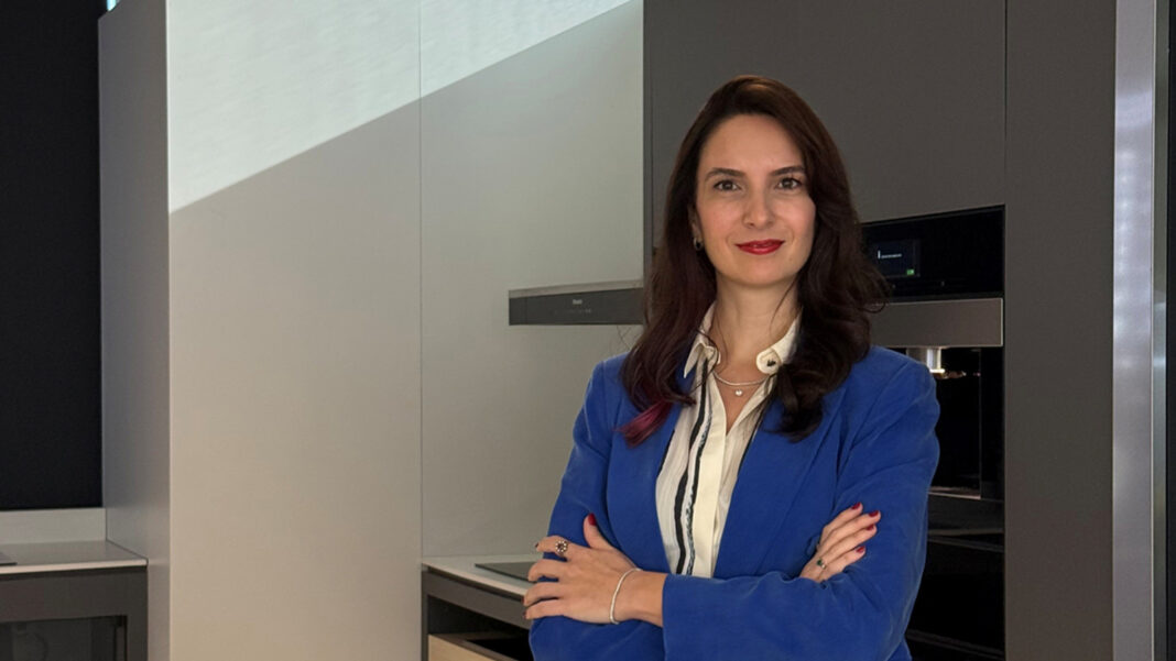 Mavi ceketli, kollarını kavuşturmuş Esra Umaçşan, ankastre mutfak cihazlarının önünde poz veriyor