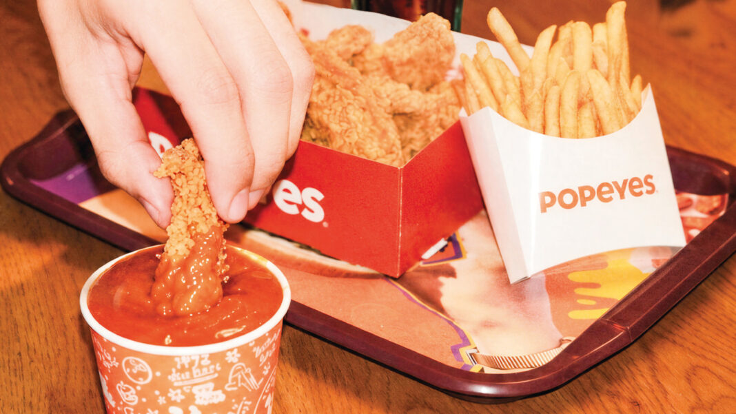 Popeyes tepsisinde patates kızartması ve kızarmış tavuk; bir el tavuk parçasını sosa batırıyor