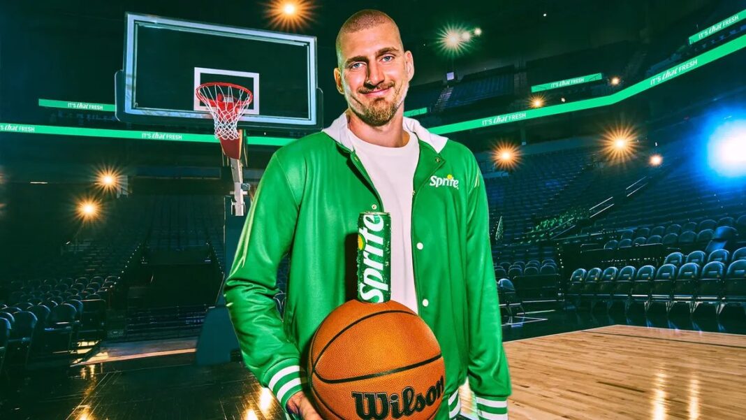 Işıklı bir basketbol salonunda yeşil Sprite ceketi giymiş, elinde Wilson basketbol topu tutan Nikola Jokić