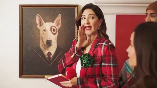 Target’ın yılbaşı reklamında ekose ceketli D’Arcy Carden, arka planda Target’ın köpek maskotu Bullseye’ın portresiyle konuşurken